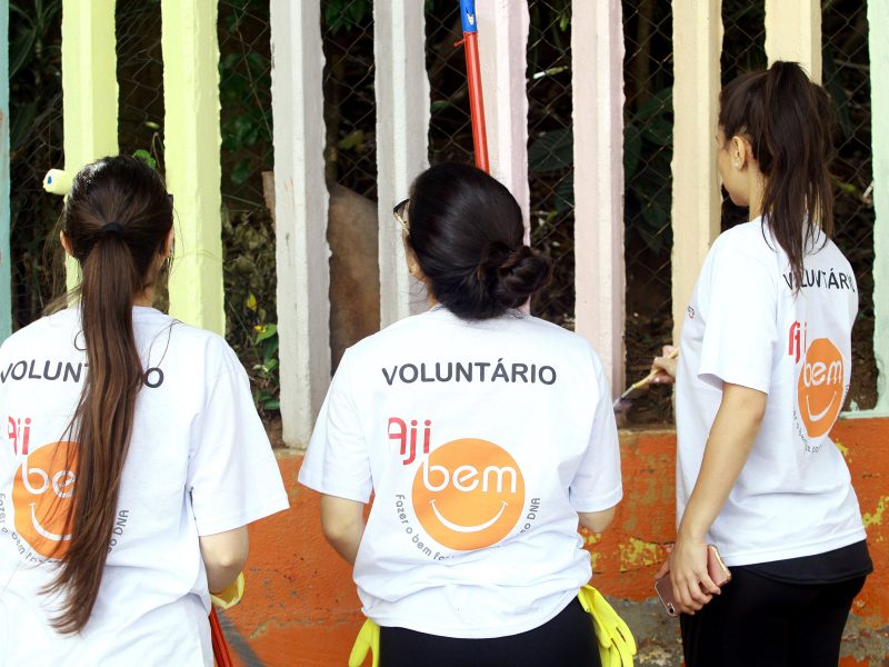 Três voluntárias estão olhando para cima, de costas, com camisetas AJIBEM